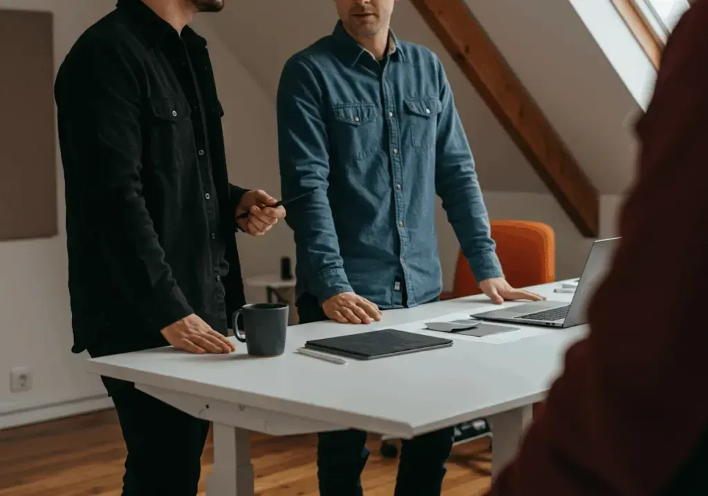 Tre personer står vid ett vitt bord med en dator, en mugg och en platta