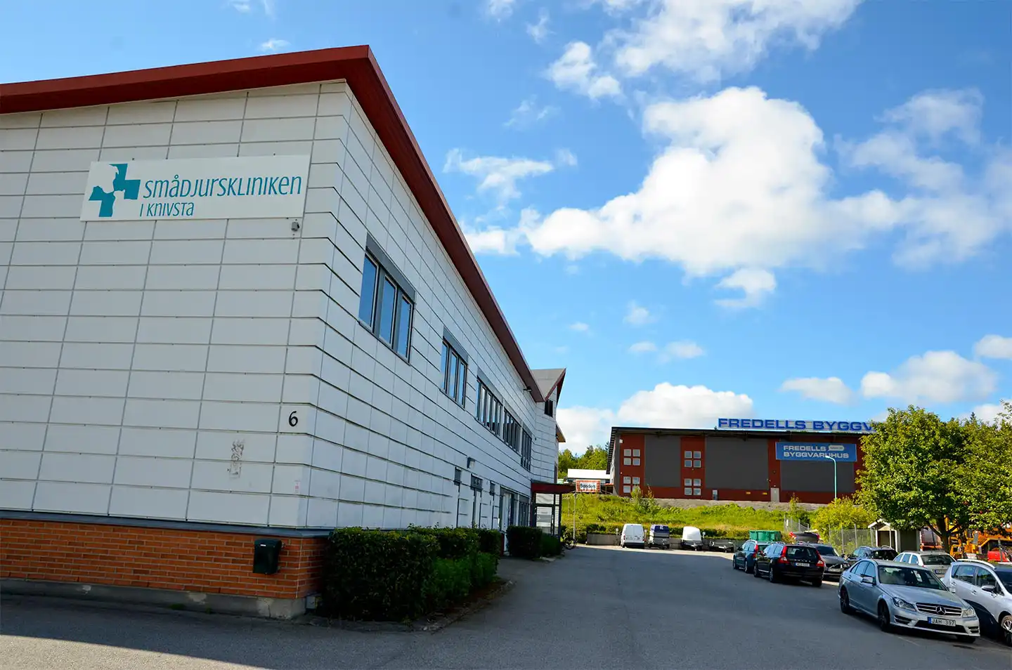 Entré till "Salong 13 Frisör" med rosa blommor i kruka. Grå byggnad i bakgrunden