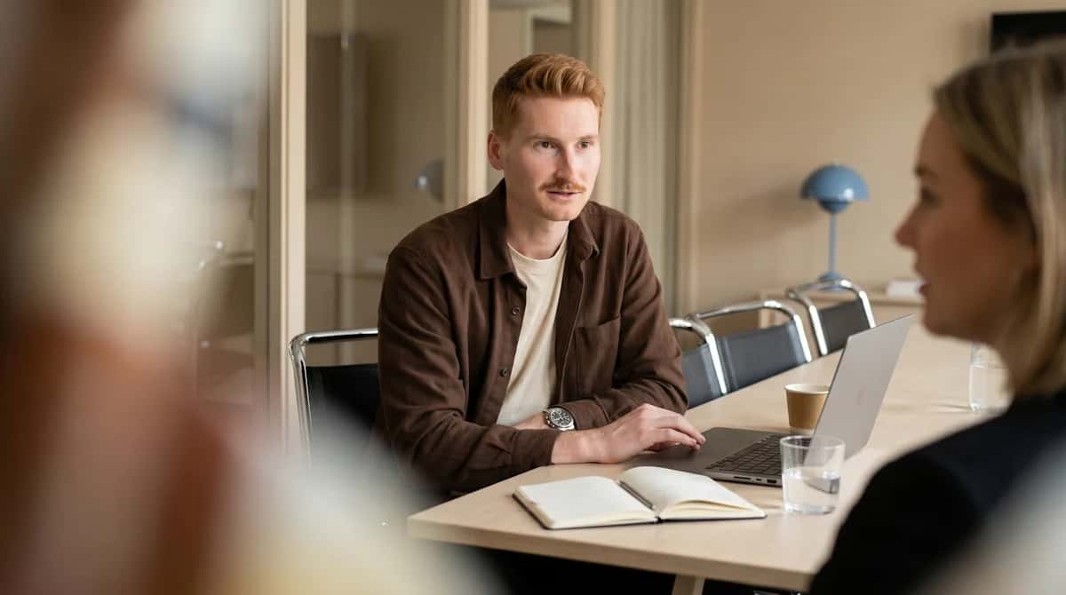 Rödhårig man med mustasch sitter vid ett bord med laptop, anteckningsbok och dryck, vänd mot kvinna i förgrunden