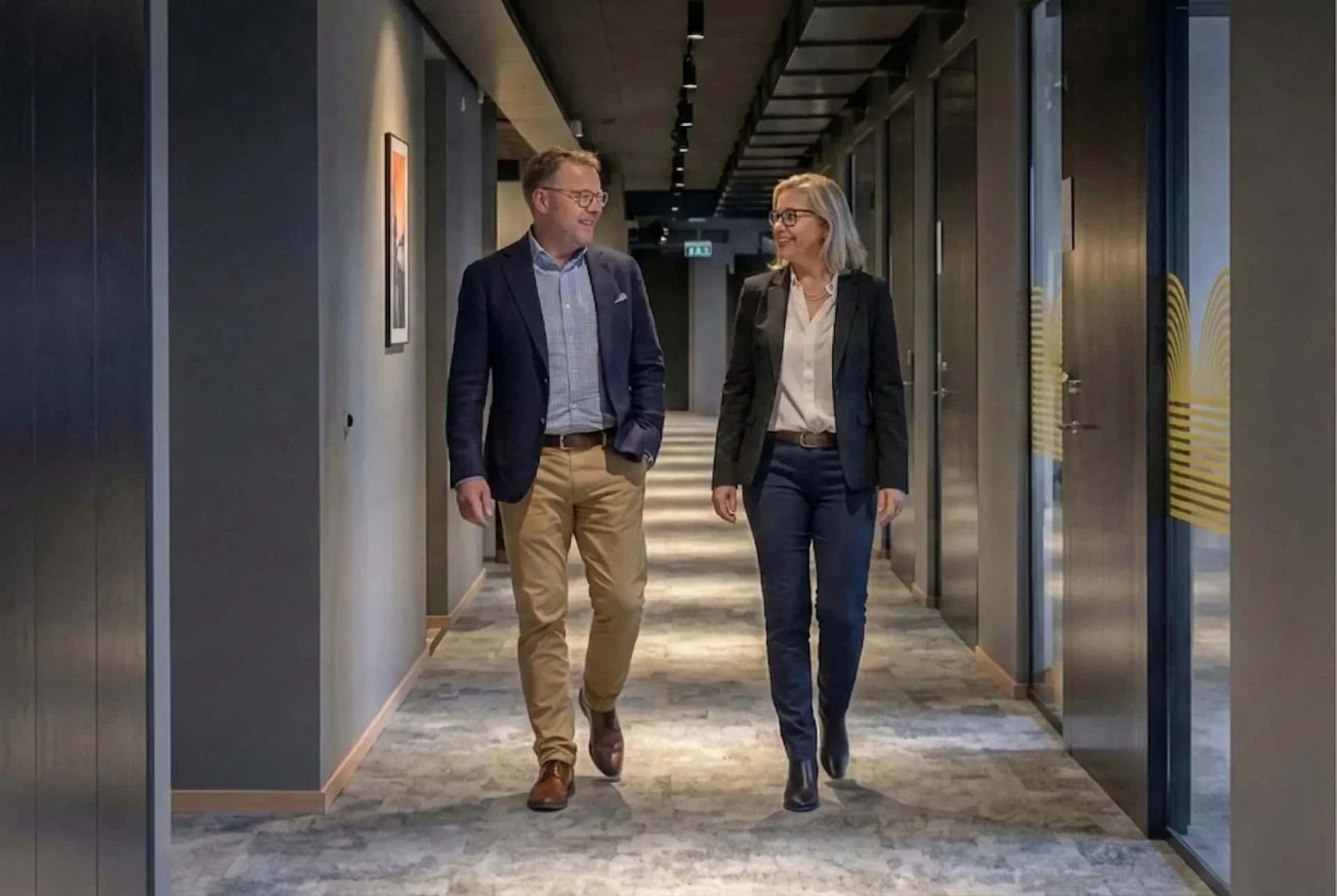 En man och en kvinna i affärskläder går i en modern kontorskorridor och ler mot varandra